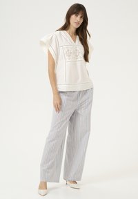 Camicia di cotone bianca con scollo a V e dettagli ricamati, abbinata a pantaloni larghi a righe verticali blu e bianchi. Tacchi beige a punta.