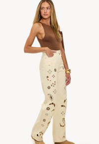 Jeans de pierna ancha color crema con patrones de flores recortados. Combinados con una blusa marrón de un solo hombro. Textura suave. Diseño elegante.