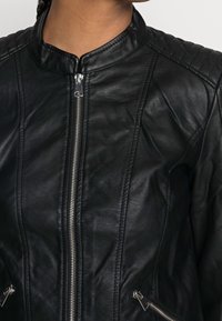Primo piano di una giacca in finta pelle nera con pannelli imbottiti sulle spalle, chiusura frontale con zip e tasche laterali con zip indossata da una persona.