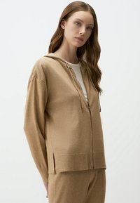 Tan hoodie met een rits aan de voorkant, lange mouwen en een ruime capuchon. Zachte gebreide stof, met zijslits en ritsdetails in goudkleur.
