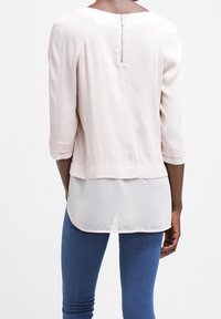 Ljust rosa blus med en mjuk textur, 3/4-ärmar och dragkedja bak. Lager på en vit, längre undertröja.