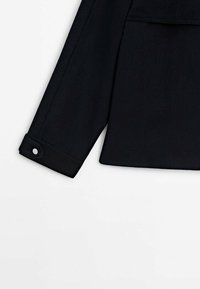 Une manche de veste noire avec un poignet boutoné. Le tissu semble lisse, avec un design ajusté et une texture subtile. Présente une coupe droite.