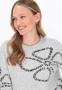 Grauer Pullover mit einem schwarzen Blumenmuster, verziert mit Perlenakzenten. Weiches Material und Rundhalsausschnitt. Lange Ärmel sichtbar.