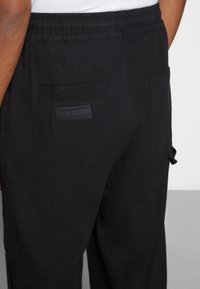 Svarta byxor i mjukt tyg med elastisk midja, två bakfickor och en liten etikett vid den högra fickan.