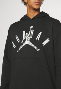 Czarna bluza z kapturem z kieszenią typu kangur. Na przodzie duże szaro-białe logo Jumpman oraz napis "JORDAN" pogrubioną czcionką na wysokości klatki piersiowej.