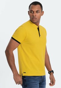 Gul polo-shirt med krave, sort knaplukning og sort kant på ærmerne. Har sideslidser og et lille logomerke ved kanten.