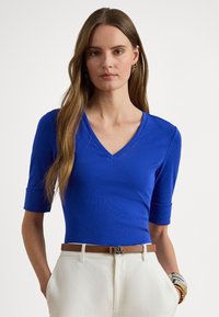 Žena oblečená v kráľovsky modrom tričku s výstrihom do V a rukávmi po lakte, biele vysoké nohavice, hnedý opasok a náramky na pravej ruke.