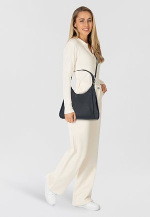 Schwarze Lederhandtasche mit dreieckiger Form, versehen mit einer strukturierten Oberfläche und einem kurzen Tragegurt. Das Modell trägt ein weißes Outfit und Sneakers.