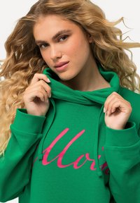 Sudadera verde con una textura suave, que presenta un diseño en script rosa en el frente, capucha con cordón y puños de canalé. Vista en primer plano.