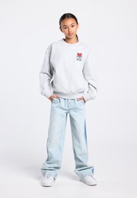 Sweat-shirt gris clair avec un petit graphique de cœur rouge, assorti à un jean bleu clair à jambes larges avec des panneaux latéraux en denim. Baskets blanches.