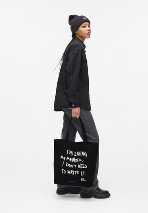 Person in black beanie, oversized black shirt, gray jeans, and platform boots carrying black tote with white text quote.