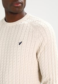 Suéter de punto de cable en color beige claro con un cuello acanalado, que presenta un pequeño logo azul marino bordado en el pecho y un detalle de tela texturizada.
