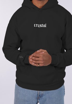 Person iført en sort hættetrøje med hvid teksten "crystal" på brystet og et lille krystalikon på venstre ærmekant, hænder foldet foran.