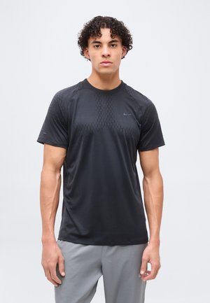 Ung mand med krøllet hår iført en sort kortærmet sportstrøje og grå bukser, stående mod en ensfarvet lys baggrund.