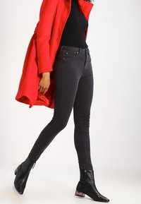 Jean skinny noir à taille haute, avec des détails de couture. Associé à un haut noir ajusté et un long manteau rouge, complété par des bottines noires.