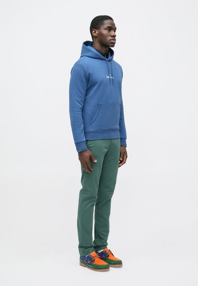 Άνδρας που στέκεται φορώντας μπλε φούτερ με κουκούλα, πράσινο παντελόνι και πολύχρωμα παπούτσια με κορδόνια σε πορτοκαλί, πράσινο και μπλε, μπροστά σε ένα απλό φωτεινό φόντο.