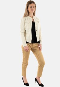 Giacca button-up beige chiaro con due tasche sul petto, maglietta nera sotto, pantaloni adatti color sabbia e scarpe con tacco nere. Design semplice, senza fantasie.