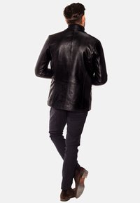 Chaqueta de cuero negra con un corte entallado, que presenta un cuello alto y detalles de costura sutiles, combinada con pantalones oscuros y zapatos marrones.
