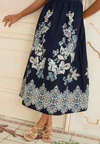 Gonna floreale blu navy con motivi bianchi e blu, caratterizzata da una vita arricciata e un dettaglio sul fondo che ricorda il pizzo. Abbinata a sandali con cinturini.