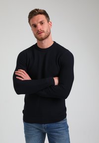 Granatowy sweter z prążkowanej dzianiny, okrągłym dekoltem i długimi rękawami, zestawiony z jasnoniebieskimi dżinsami, prezentowany przez osobę z skrzyżowanymi ramionami.