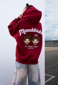 Persona che indossa un felpa bordeaux con la scritta "Monchhichi" e personaggi dei cartoni animati sul retro, abbinata a jeans azzurri a fantasia.