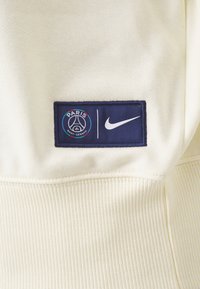 Tela beige con un parche azul marino que muestra el logo de Paris Saint-Germain y el swoosh blanco de Nike uno al lado del otro.