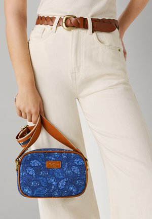 Persona con pantalones color crema y un cinturón trenzado marrón, llevando un bolso cruzado azul con patrón y correa marrón.