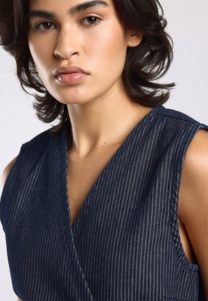 Young person with medium-length dark hair wearing a sleeveless, dark blue pinstriped top, looking slightly to the side.