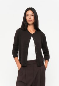 Mørkebrun cardigan laget av mykt materiale, med rund hals, knapp front og ribbede mansjetter. Bæres over en hvit topp.