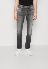 Persona con jeans ajustados negros desgastados y ligeros desgastes, y zapatillas blancas, de pie contra un fondo blanco liso.