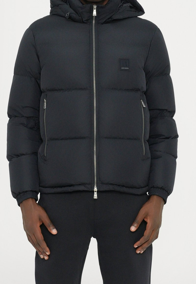 Veste matelassée noire avec capuche, dotée d'une fermeture éclair, de poches latérales et de poignets élastiques. Texture lisse avec un motif matelassé.