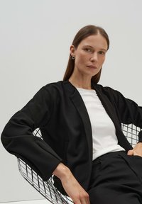 Blazer noir au design ajusté, avec des manches longues, une ouverture frontale et deux poches, porté sur un haut blanc, en contraste avec une chaise en fil.