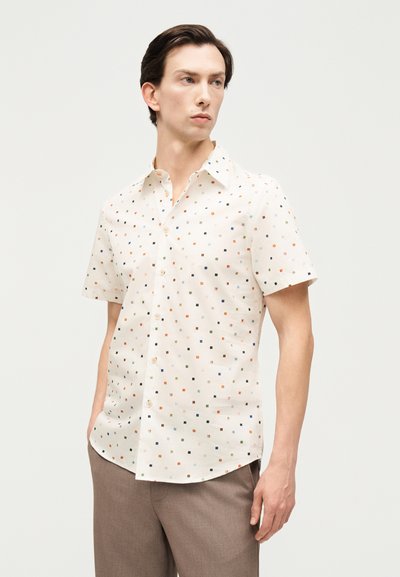 Νεαρός άνδρας που φοράει λευκό πουκάμισο με κοντά μανίκια και πολύχρωμα μικρά τετράγωνα σχέδια, μαζί με καφέ παντελόνι, στέκεται μπροστά σε απλό φόντο.