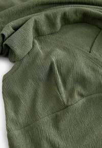 Gros plan sur un tissu texturé vert olive avec des détails froncés et des manches à volants sur un vêtement ajusté.