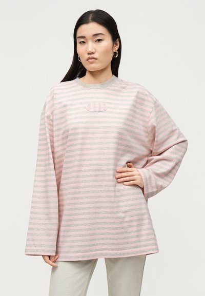 Νεαρή γυναίκα με μακριά σκούρα μαλλιά φοράει ριγέ ροζ και γκρι φαρδιά πουλόβερ και ανοιχτόχρωμο παντελόνι, ποζάροντας με το χέρι στη μέση.