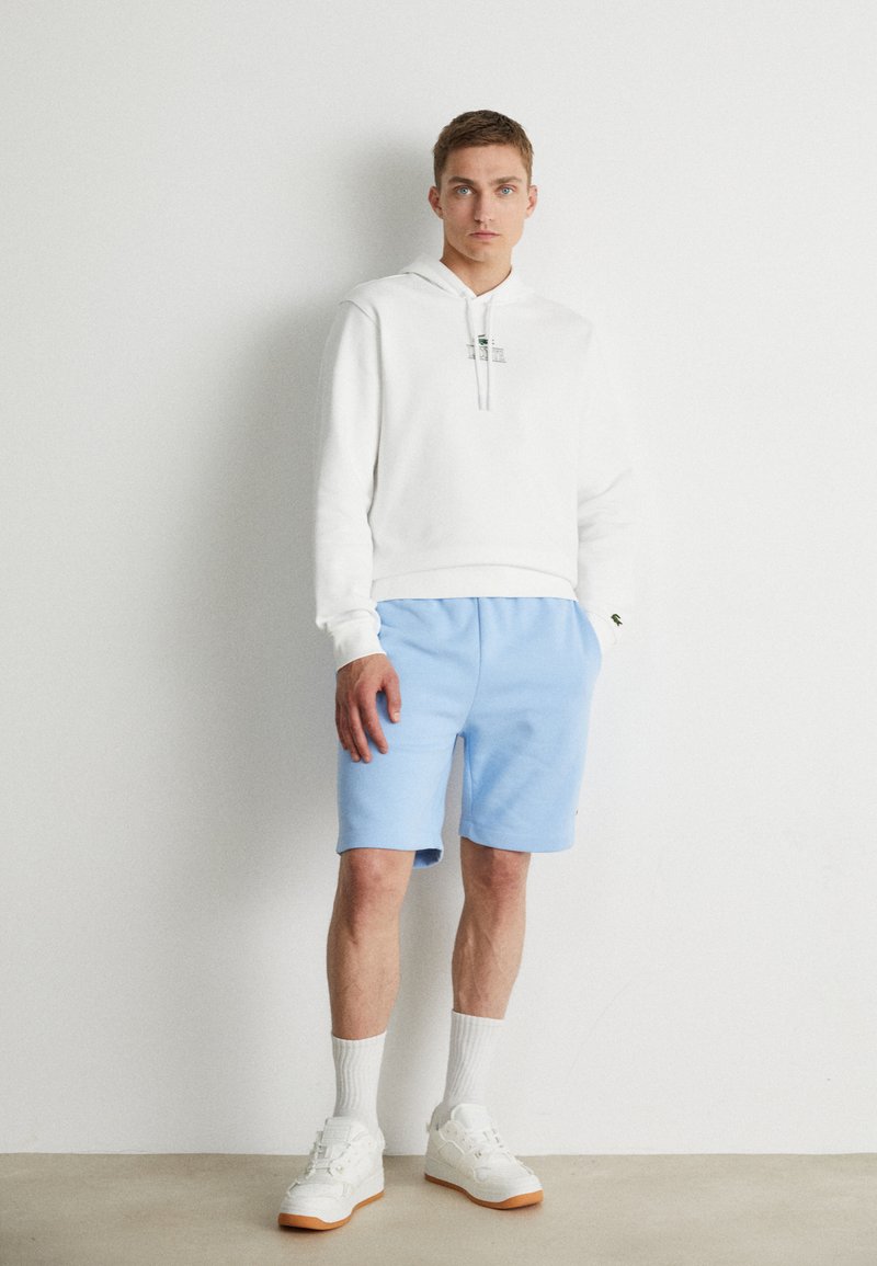 Sweatshirt à capuche blanc, short bleu clair et baskets blanches avec semelles en caoutchouc. Tenue décontractée avec des poignets côtelés et des accents brodés.