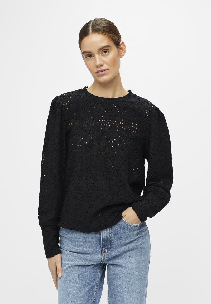 Blouse noire à manches longues avec un motif en œillets, col rond et épaules froncées, associée à un jean bleu clair.