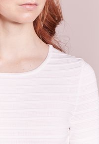 Haut blanc texturé avec des nervures horizontales, col rond, coupe ajustée et tissu lisse. Vue en gros plan mettant l'accent sur le col et la zone des épaules.