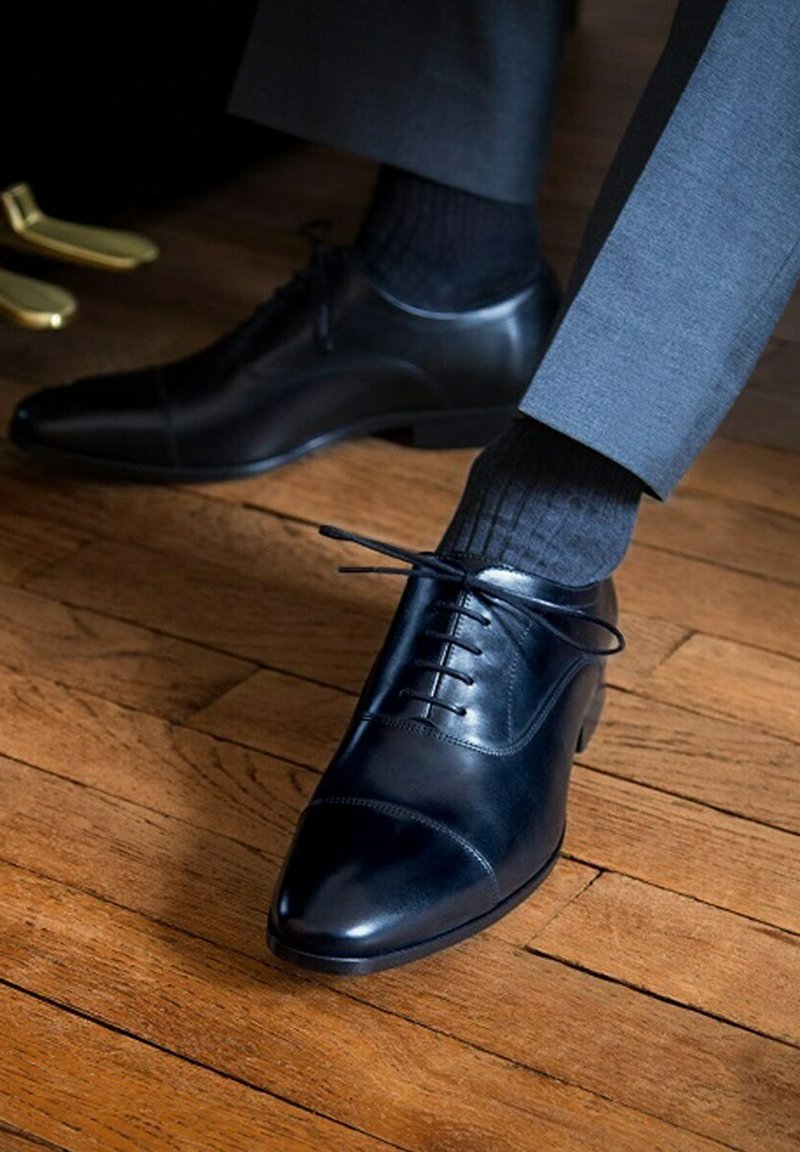 Zwarte leren dresschoenen met een gladde, gepolijste afwerking, ronde neus en vetersluiting. Gecombineerd met donkere geribbelde sokken en grijze broek.