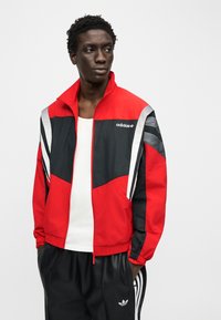 Νεαρός άνδρας που φοράει κόκκινη και μαύρη ζακέτα φόρμας Adidas πάνω από λευκό αμάνικο και μαύρο παντελόνι φόρμας Adidas, με τα χέρια στις τσέπες.