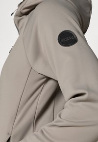 Beige mīkstās apvalka jakas ar kapuci, teksturētu audumu un melnu apļveida logotipa uzlīmi augšējā rokā. Redzamas kabatas ar rāvējslēdzēju.