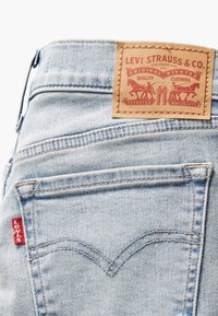 Vaqueros de mezclilla azul claro con una etiqueta marrón que presenta un logo en la parte posterior de la cintura. Cuenta con costuras rojas y un diseño de bolsillo trasero distintivo.