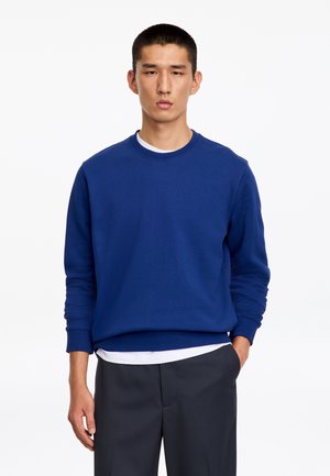 Homme aux cheveux courts portant un sweat-shirt bleu à col ras du cou par-dessus une chemise blanche et un pantalon foncé, debout avec une main dans la poche sur fond blanc.