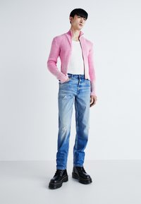 Šviesiai rožinė banguota zip-up megztinis virš balto tank top'o, derinamas su suplėšytais šviesiai mėlynais džinsais ir juodais masyviais bateliais su raišteliais.