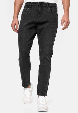 Homme portant un jean noir slim et des baskets blanches, debout devant un fond clair uni.