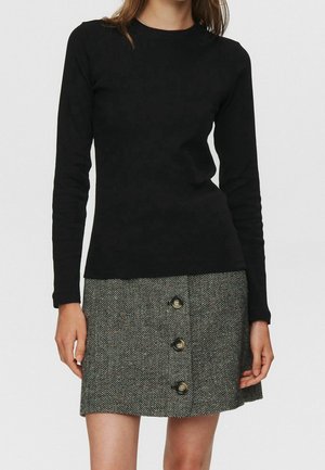 Kobieta ubrana w czarną bluzkę z długim rękawem i szarą spódnicę jodełkową z trzema guzikami z przodu, stojąca na tle gładkiej białej ściany.