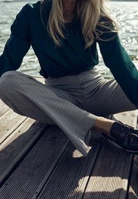 Haut à manches longues vert foncé, pantalon large à carreaux noir et blanc, et chaussures foncées. Position assise sur une surface en bois près de l'eau.