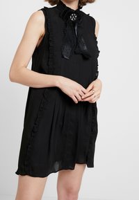 Černé bezramínkové šaty s volánkovými lemy, vysokým límcem a detaily s mašlí. Mají strukturovanou tkaninu a dekorativní brož.