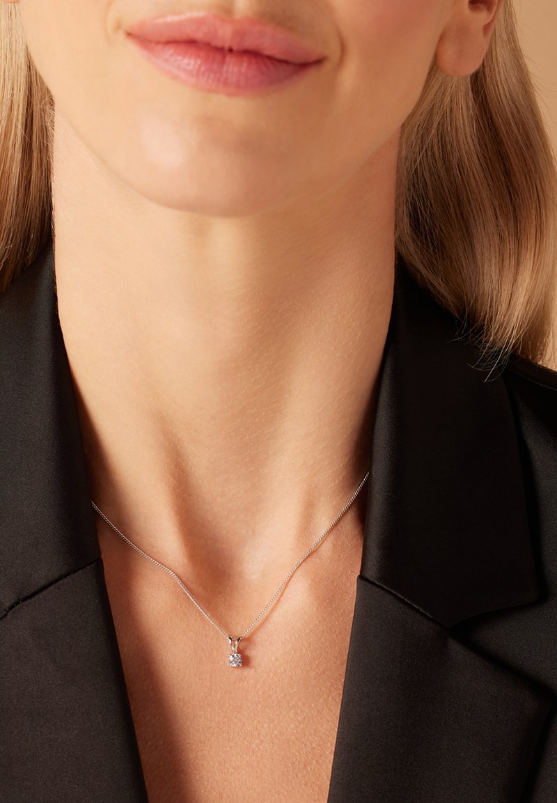 Zilveren ketting met een kleine, transparante edelsteenhanger, gedragen tegen een blootgestelde nek, aangevuld met een zwarte blazer.