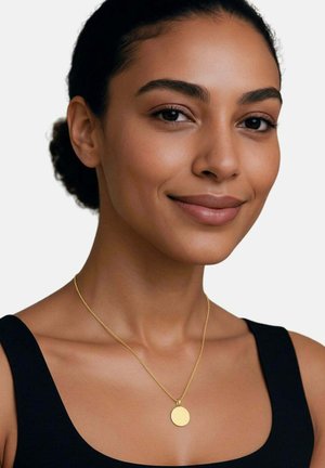 Junge Frau mit glatter Haut und zurückgebundenem dunklem Haar, die eine goldene runde Anhängerhalskette und ein schwarzes ärmelloses Oberteil trägt und sanft lächelt.